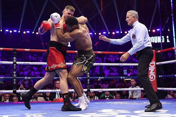 Anthony Joshua Vs Prenga Trybal News Two boxers grapple in the ring as a referee watches one in red trunks with red gloves the other in camouflage trunks with white gloves crowd visible in the background trybalnews