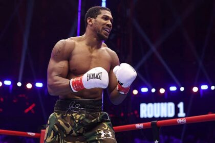 Male boxer in camouflage shorts and white Everlast gloves in a boxing ring, fists up during Round 1 with bright arena lights in the background.