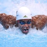 Swimmer wearing a white cap and blue goggles performs the butterfly stroke breaking through the blue pool water with splashes around  trybalnews