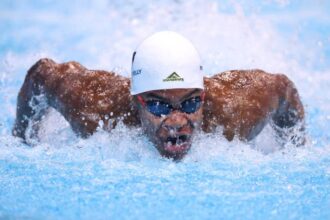Swimmer wearing a white cap and blue goggles performs the butterfly stroke, breaking through the blue pool water with splashes around.