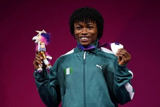 Young athlete in a teal PUMA jacket stands against a magenta background, proudly holding a small trophy with a plush toy in one hand and medals in the other, smiling at the camera.