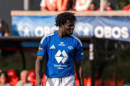 Male soccer player in blue jersey and white shorts walking on a green pitch during a game, with spectators in the background.