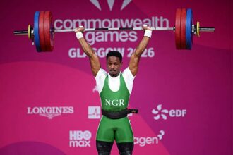 Nigerian weightlifter in green lifts a loaded barbell overhead at the Commonwealth Games stage (2016).