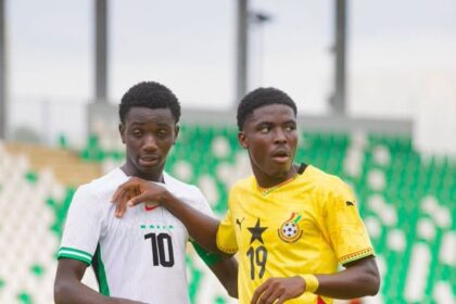 Two male soccer players on a field, one in white with the number 10 and green trim, the other in yellow with the number 19, standing close in a stadium.