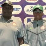 Two men pose for a photo at a sports event backdrop with sponsor logos both wear gray tops one with a cap and cross necklace the other in a green cap and zip jacket with a crest on the chest  trybalnews