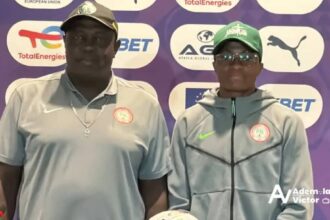 Two men pose for a photo at a sports event backdrop with sponsor logos; both wear gray tops, one with a cap and cross necklace, the other in a green cap and zip jacket with a crest on the chest.
