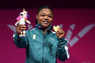 Female athlete on a podium smiling, holding a gold medal in one hand and a plush unicorn trophy in the other, wearing a teal jacket with an Irish flag patch