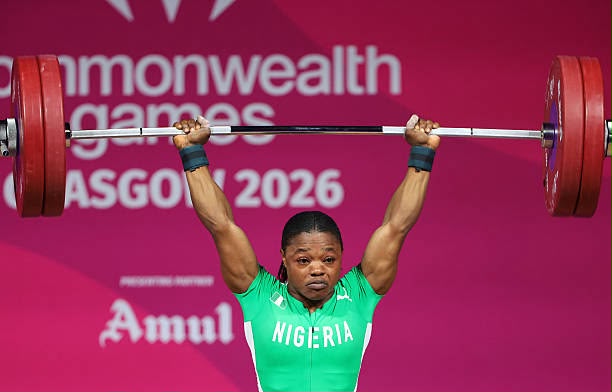 Glasgow 2026 Trybal News Nigerian weightlifter lifting a heavy barbell overhead at the Commonwealth Games 2026 wearing a green jersey against a pink backdrop trybalnews