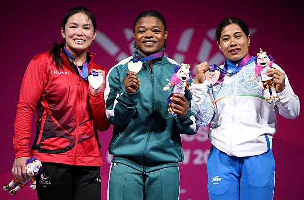 Glasgow 2026 Trybal News Three female athletes pose on a podium smiling and displaying medals and mascot figurines trybalnews