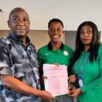 Three people stand indoors as they hand a pink certificate celebrating an achievement  trybalnews