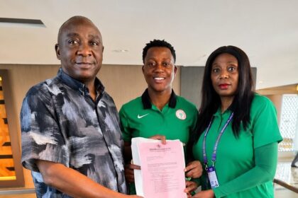 Three people stand indoors as they hand a pink certificate, celebrating an achievement.