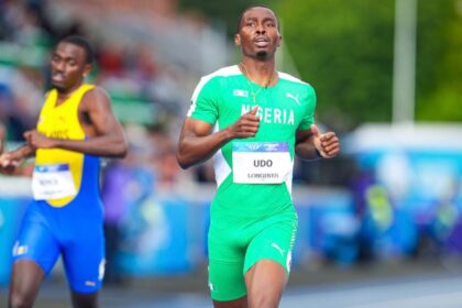 Two male sprinters race on a track; foreground runner in green Nigeria kit with bib ‘UDO LONGINES’.
