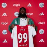 Athlete with dreadlocks holds up a red and white jersey labeled TOLU with the number 99 in front of a red sponsor wall  trybalnews