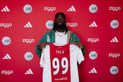 Athlete with dreadlocks holds up a red-and-white jersey labeled 'TOLU' with the number 99 in front of a red sponsor wall.