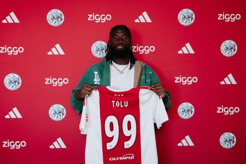 Athlete with dreadlocks holds up a red and white jersey labeled TOLU with the number 99 in front of a red sponsor wall  trybalnews
