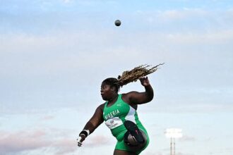 Nigerian female hammer thrower in a green uniform mid-throw with the ball arcing overhead against a blue sky backdrop.