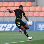 Soccer player in a black kit with neon green accents kicking a white ball on a green pitch orange stadium seats in the background  trybalnews