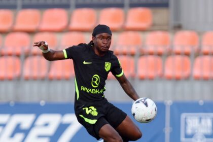 Soccer player in a black kit with neon green accents kicking a white ball on a green pitch, orange stadium seats in the background.