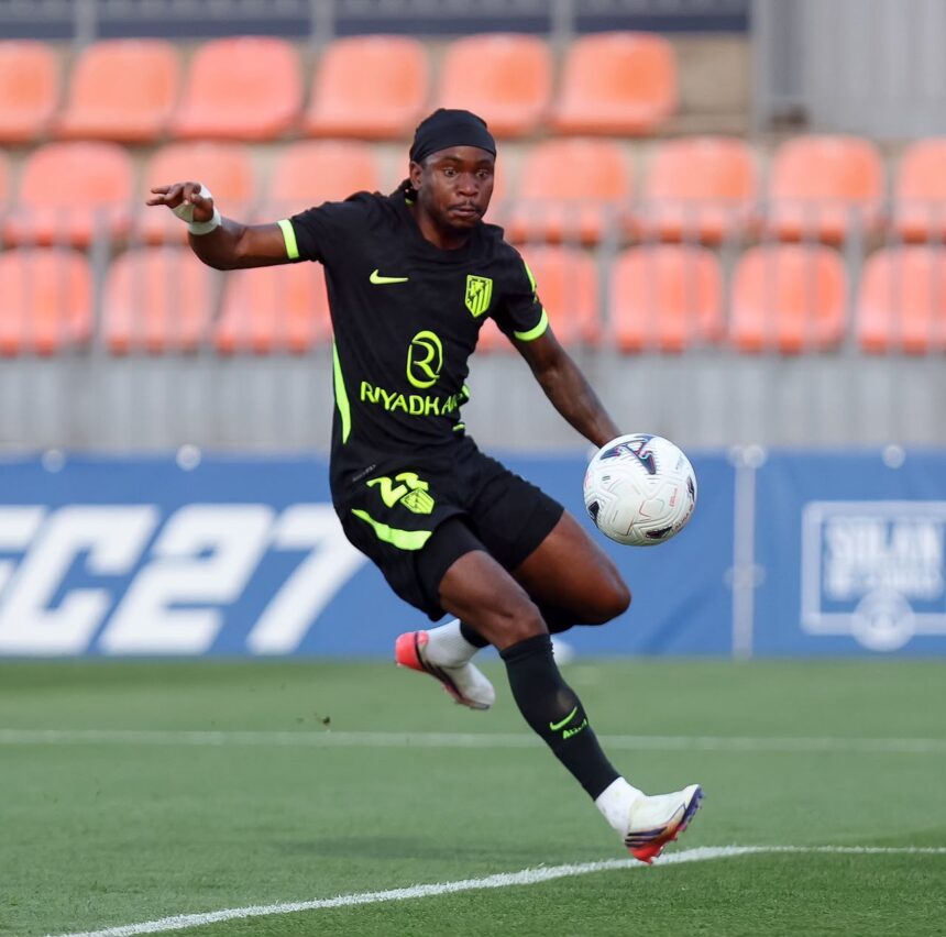 Soccer player in a black kit with neon green accents kicking a white ball on a green pitch orange stadium seats in the background  trybalnews