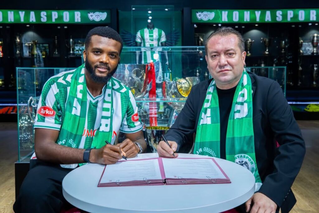Chidozie Awaziem Trybal News Two men in green and white team scarves sign a contract at a small round table with a trophy filled display behind them trybalnews