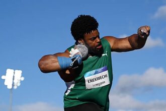 Male track and field athlete in a green uniform mid-throw, bib ENEKWECHI LONGINES, against a blue sky background.