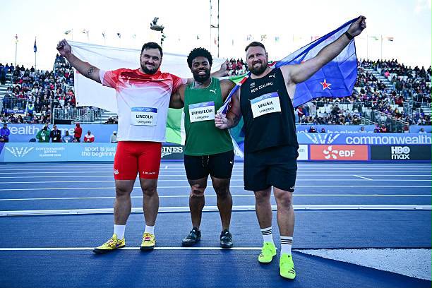 Glasgow 2026 Trybal News Three male sprinters on a blue track celebrate with arms around each other holding flags as a cheering crowd watches in the stands trybalnews
