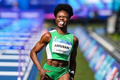 Female sprinter in a bright green uniform celebrating at a track event, tongue out, bib reads AMUSAN.