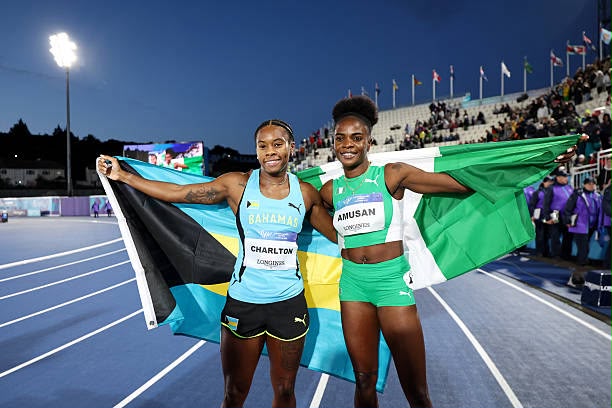 Glasgow 2026 Trybal News Two female sprinters celebrate with national flags Bahamas and Nigeria after a race smiling on the blue track with a stadium crowd behind them trybalnews