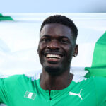 Smiling Black male soccer player in a green jersey with a green and white flag behind him arms outstretched in celebration the image overall suggests victory  trybalnews