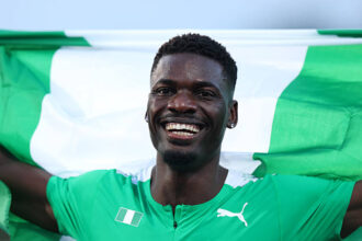 Smiling Black male soccer player in a green jersey with a green-and-white flag behind him, arms outstretched in celebration the image overall suggests victory.