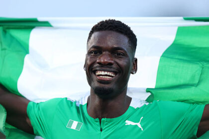 Smiling Black male soccer player in a green jersey with a green-and-white flag behind him, arms outstretched in celebration the image overall suggests victory.