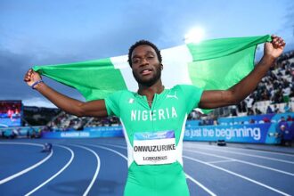 Nigerian sprinter on the track celebrates with a green Nigerian flag raised behind him, crowded stadium in the background.