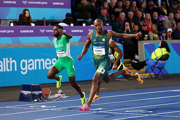 Glasgow 2026 Trybal News Two male sprinters in green uniforms race on a blue track at a stadium with spectators and banners in the background trybalnews