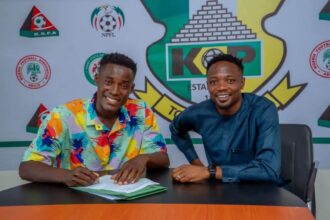 Zayyad Musa, wearing a colorful multi-colored shirt, signs his contract at a table while seated next to Kano Pillars Club Ambassador Ahmed Musa in a blue traditional outfit. Both are smiling. The Kano Pillars crest and NPFL logos are visible on the backdrop. Photo Credit: Kano Pillars