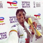 Smiling female footballer in a white jersey with number 14 holds a tall gold trophy at a Woman of the Match ceremony backdrop with sponsor logos like TotalEnergies  trybalnews