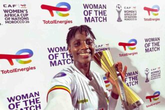 Smiling female footballer in a white jersey with number 14 holds a tall gold trophy at a 'Woman of the Match' ceremony backdrop with sponsor logos like TotalEnergies.