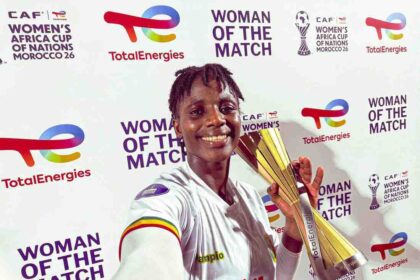 Smiling female footballer in a white jersey with number 14 holds a tall gold trophy at a 'Woman of the Match' ceremony backdrop with sponsor logos like TotalEnergies.