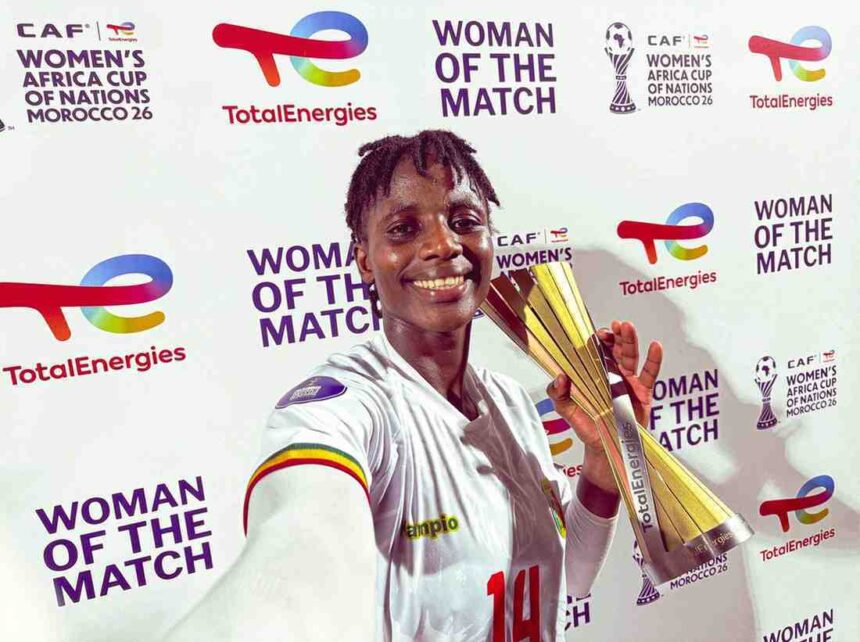 Smiling female footballer in a white jersey with number 14 holds a tall gold trophy at a Woman of the Match ceremony backdrop with sponsor logos like TotalEnergies  trybalnews