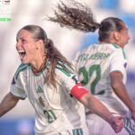 Two Algeria womens soccer players celebrate on the field the foreground player wears number 21 and a red captains armband while her teammate runs behind  trybalnews