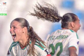 Two Algeria women’s soccer players celebrate on the field; the foreground player wears number 21 and a red captain’s armband while her teammate runs behind.
