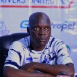 A bald man in a white and blue sports jersey sits at a press conference table with his arms crossed on the table in front of him  trybalnews