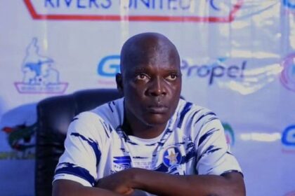 A bald man in a white and blue sports jersey sits at a press conference table with his arms crossed on the table in front of him.
