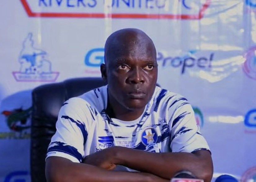 A bald man in a white and blue sports jersey sits at a press conference table with his arms crossed on the table in front of him  trybalnews