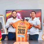 Two men in white shirts hold up an orange sports jersey labeled 2028 in a formal room setting smiling for the camera  trybalnews