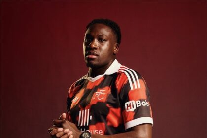 Portrait of a male soccer player wearing a red and black club jersey, hands clasped, against a dark red backdrop.