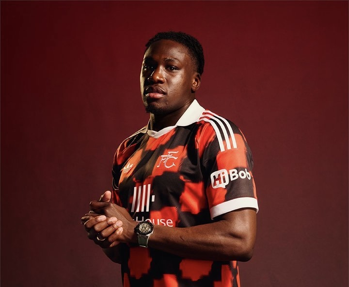 Portrait of a male soccer player wearing a red and black club jersey hands clasped against a dark red backdrop  trybalnews