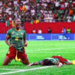 Cameroonian footballer wearing green jersey number 10 celebrates a goal on a green pitch with a fallen opponent nearby and a cheering crowd in the stands  trybalnews
