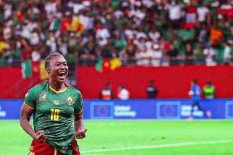 Cameroonian footballer wearing green jersey number 10 celebrates a goal on a green pitch, with a fallen opponent nearby and a cheering crowd in the stands.
