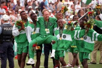 Nigeria's head coach Johannes Bonfrere is chaired in the air by his players wearing green and white tracksuits after Nigeria beat Argentina 3-2 in the Football Final at the 1996 Olympic Games in Atlanta, USA. The gold medalists smile, hold bouquets and wear gold medals around their necks while celebrating on the pitch with fans in the background.