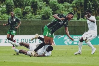 Soccer players in green and white chase the ball on a grass field; one player tumbles as others move toward the ball.